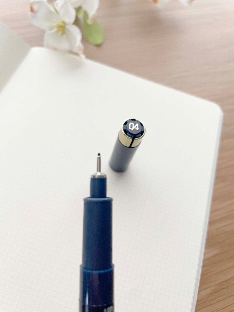 May include: A blue and silver fineliner pen with a black cap and the number "04" printed on the cap. The pen is lying on a white notebook.