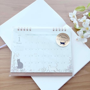 May include: A 2027 desk calendar with a spiral binding, featuring a January page with illustrations of black and white cats among flowers. A circular gold sticker on the top right displays the text 2027 CALENDAR with a black cat silhouette.