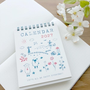 May include: A spiral-bound 2027 calendar with a white cover featuring blue and red line drawings of sheep, flowers, windmills, and a lighthouse. The text reads CALENDAR 2027 and LET'S ALL BE HAPPY EVERYDAY! The calendar is resting on a white notebook.