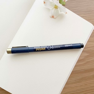 May include: A white notebook with a grid pattern, a black pen with gold accents, and white flowers on a wooden surface. The pen is labeled "Sakura Micron Pigma 04" and "Water Resistant and Fade Proof Ink."