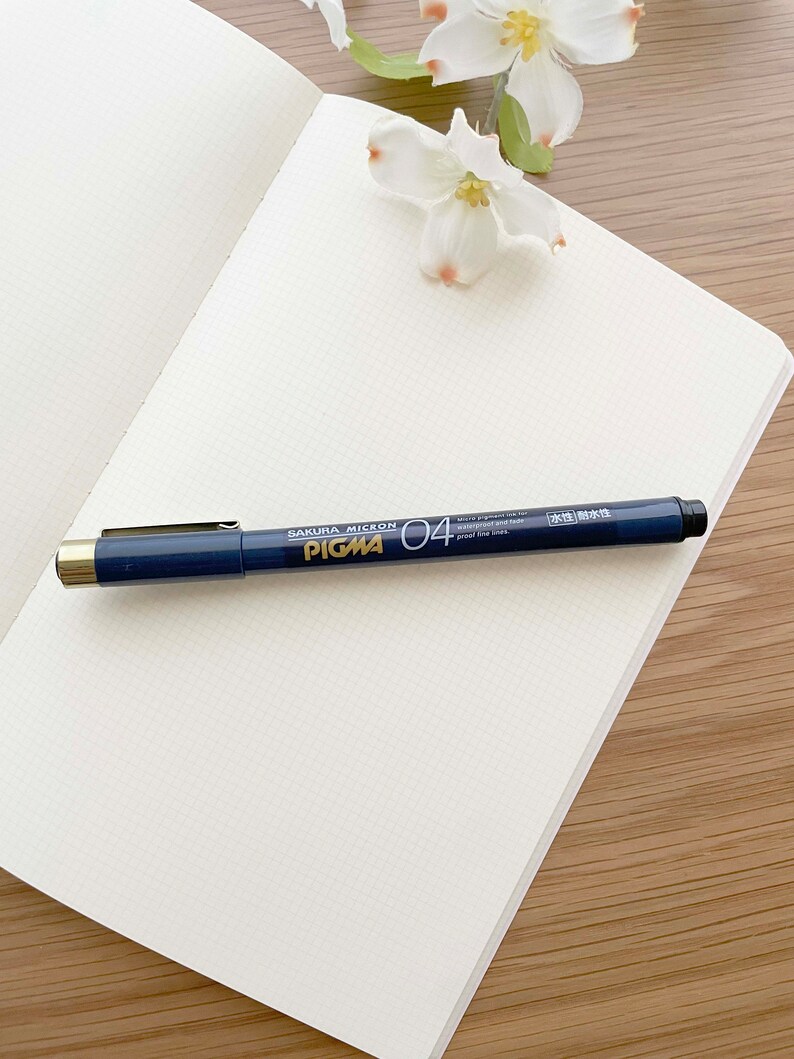 May include: A white notebook with a grid pattern, a black pen with gold accents, and white flowers on a wooden surface. The pen is labeled "Sakura Micron Pigma 04" and "Water Resistant and Fade Proof Ink."