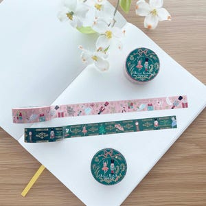 May include: Decorative washi tape rolls and strips with holiday-themed designs. One roll and strip feature a pink background with nutcrackers and gifts. Another roll and strip have a dark green background with ballerinas and Christmas trees. The tape is on a white notebook.