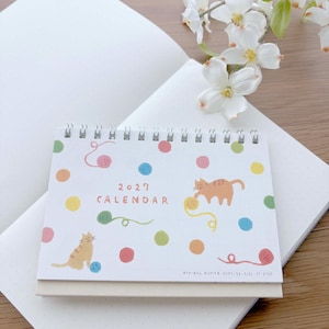 May include: A 2027 desk calendar with a white background featuring colorful polka dots, illustrations of two orange tabby cats, and yarn balls. The calendar is spiral-bound at the top and includes the text 2027 CALENDAR in a simple font.