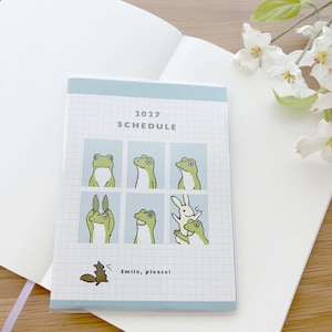 May include: A 2027 schedule planner with a grid pattern cover featuring six panels of green frog illustrations, one panel showing a white rabbit, and a small squirrel graphic at the bottom with the text "Smile, please!" printed in black.