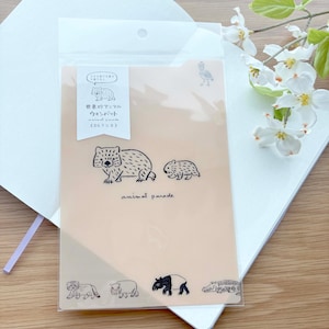 May include: A clear plastic sleeve containing a peach-colored paper with black line drawings of animals. The words "animal parade" are written below the illustrations. The sleeve is on top of a white notebook.