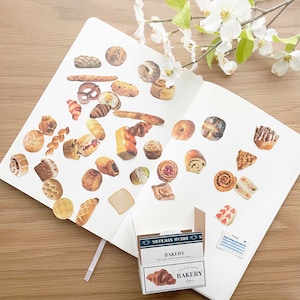 May include: A white notebook with a variety of bread stickers on the pages. The stickers are in the shape of different types of bread, including croissants, baguettes, and rolls. The notebook is open to a page with a sticker of a croissant on it. The notebook is sitting on a wooden table. The text on the box reads "Bread Stickers" and "Bakery".