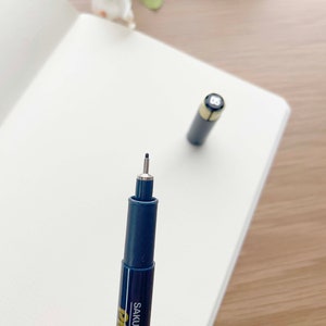 May include: A black and gold Sakura Micron Pigma pen with a fine tip. The pen is on a white surface.