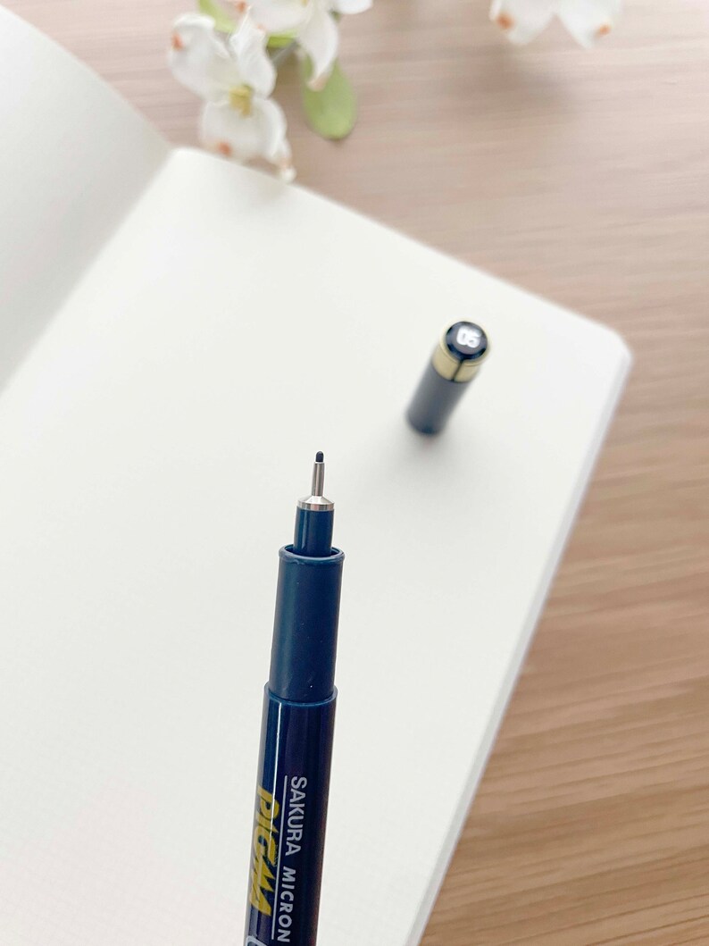May include: A black and gold Sakura Micron Pigma pen with a fine tip. The pen is on a white surface.