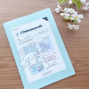 May include: A light blue folder with a grid pattern and a cartoon illustration of a dog character named Cinnamoroll. The text "Cinnamoroll" is in black and the text "A warm heart, a wagging tail, and a smile just for you!" is in black. The illustration shows the dog character with a heart, a tail, and a smile.