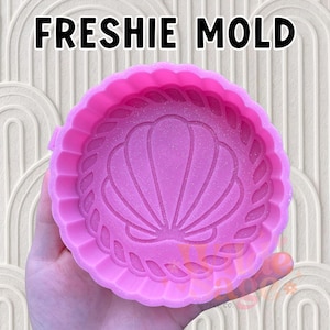 May include: A pink silicone mold with a seashell design and the text "FRESHIE MOLD" in black. The mold has a scalloped edge and a textured surface. The seashell design is in the center of the mold.