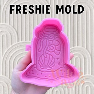 May include: A pink silicone mold with the text "FRESHIE MOLD" at the top. The mold features a seashell design inside a jar shape. The mold is held in a hand.