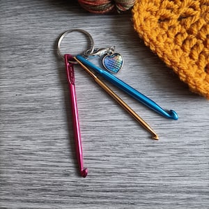 May include: A keyring featuring three miniature metal crochet hooks in metallic pink, gold, and blue, attached to a silver ring. A small heart-shaped charm with a blue and green iridescent scale pattern hangs from the ring alongside the hooks.