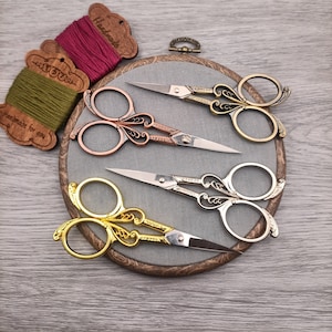 May include: Three pairs of decorative embroidery scissors with ornate handles. The scissors are arranged on a round, wooden embroidery hoop with a grey fabric background. The scissors are gold, silver, and copper in colour.