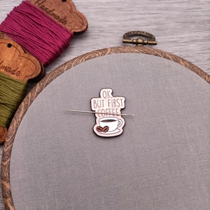 Könnte beinhalten: Ein brauner Holz-Stickrahmen mit einem grauen Leinenstoff im Inneren. Eine goldfarbene Emaille-Nadel mit einer Kaffeetasse und dem Text "OK, BUT FIRST COFFEE" ist auf den Stoff gesteckt.