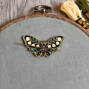 Könnte beinhalten: Eine goldfarbene Schmetterlingsnadel mit schwarzem, grünem und weißem Emaille. Der Schmetterling hat einen Halbmond und Sterne auf seinen Flügeln. Die Nadel befindet sich auf einem grauen Stoffhintergrund.