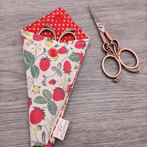 May include: Decorative scissors with a copper-coloured handle and a strawberry-patterned fabric case. The case is red and white with a red polka dot lining. The scissors are next to the case on a wooden surface. The case has the text "Botanical Craft Studios".