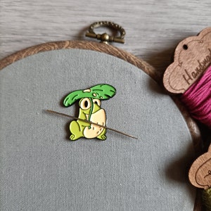 Puede incluir: Pin de esmalte de rana verde con una hoja en la cabeza. El pin está sobre un fondo de tela gris con una aguja e hilo.