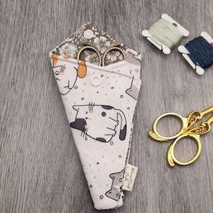 May include: A triangular fabric scissor sheath featuring a white background with a black and grey cat pattern, secured by a white snap button. The interior lining has a grey floral print. A pair of gold-toned metal scissors rests nearby.