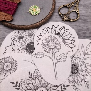 May include: A set of four white fabric embroidery patterns featuring black line drawings of sunflowers. The patterns are arranged in a stack, with the top pattern showing a single sunflower with leaves. The patterns are designed for embroidery projects.