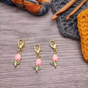 May include: Three gold-colored tulip stitch markers with pink petals and green leaves. The markers are attached to clasps. A skein of blue, orange, and brown yarn, along with crochet hooks and yarn samples, are in the background.