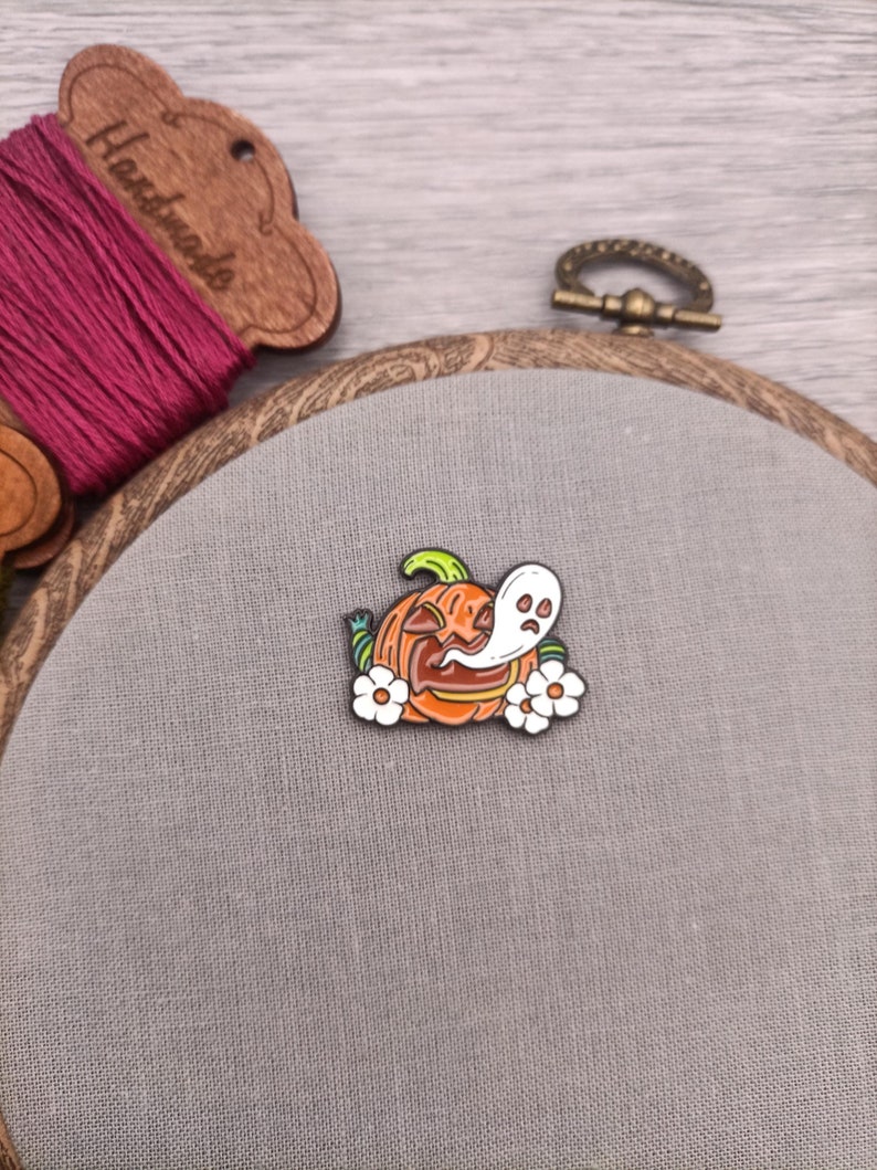 May include: A metal enamel pin featuring a cartoon pumpkin with a ghost inside. The pumpkin is orange with a green stem and black eyes and mouth. The ghost is white with black eyes and a small mouth. There are three white flowers around the pumpkin.