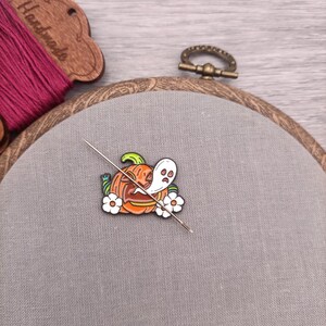 May include: A close-up of a needle and thread going through a Halloween-themed enamel pin. The pin features a jack-o'-lantern with a ghost inside and white flowers.