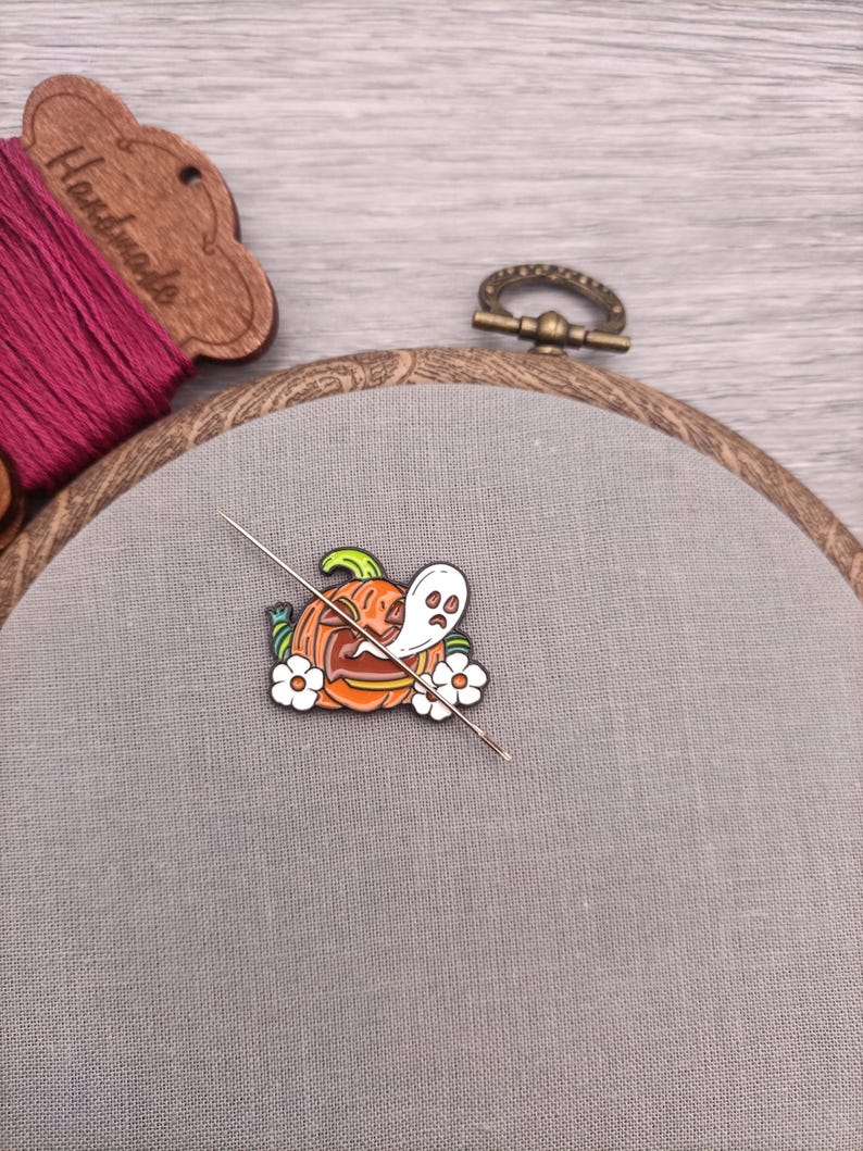 May include: A close-up of a needle and thread going through a Halloween-themed enamel pin. The pin features a jack-o'-lantern with a ghost inside and white flowers.