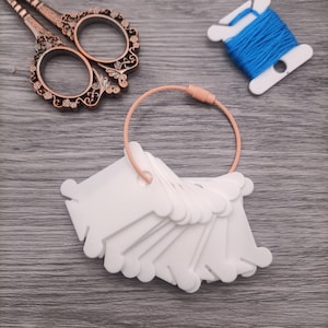 Könnte beinhalten: Ein Satz weißer Kunststoff-Fadenspulen mit einer Kupferspule. Die Spulen sind übereinander gestapelt. Eine Schere mit Verzierungen und eine Spule blauen Fadens sind ebenfalls im Bild.