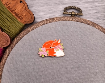 May include: A gold-toned enamel pin featuring a curled-up orange fox with a white tail tip and pink flowers. The pin is on a gray fabric background.