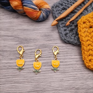 May include: Three yellow tulip-shaped stitch markers with gold-colored clasps. The markers are set against a backdrop of crochet hooks, yarn, and crocheted fabric. The yarn is a mix of blue, orange, and brown.