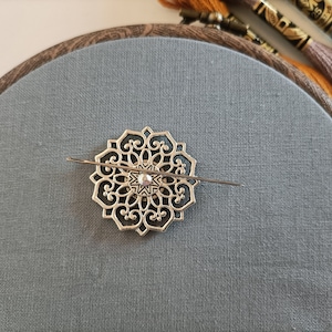 May include: A gold-coloured metal embroidery hoop with a decorative filigree design. The hoop is on a grey fabric background.