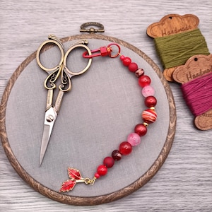 Può includere: Forbici da ricamo in stile vintage con manici ornati appoggiate su un telaio ricamato rivestito in tessuto grigio. Un portafob a forbice con perline rosse con un ciondolo a forma di foglia è appeso alle forbici.