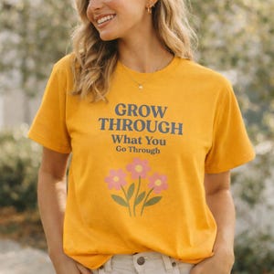 Peut inclure: T-shirt jaune moutarde avec le texte "GROW THROUGH What You Go Through" en bleu marine. En dessous du texte, trois fleurs roses avec des tiges et des feuilles vertes. Le t-shirt a des manches courtes.