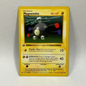 May include: A yellow-bordered Magnemite Pokémon card. The card features a black, silver, red, and blue Magnemite illustration. The card text includes "Thunder Wave" and "Selfdestruct" attack descriptions. The card is 1 foot long and weighs 13 lbs.