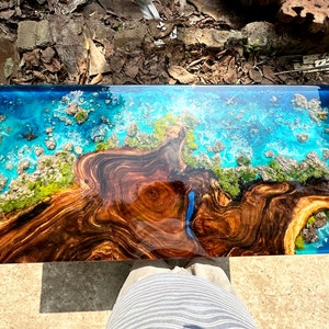 Puede incluir: Una mesa de centro rectangular con una base de madera oscura y una tapa de resina epoxi azul. La tapa de resina presenta un dise&ntilde;o de un oc&eacute;ano azul con olas blancas e islas verdes. La mesa mide aproximadamente 120 cm de largo y 60 cm de ancho.