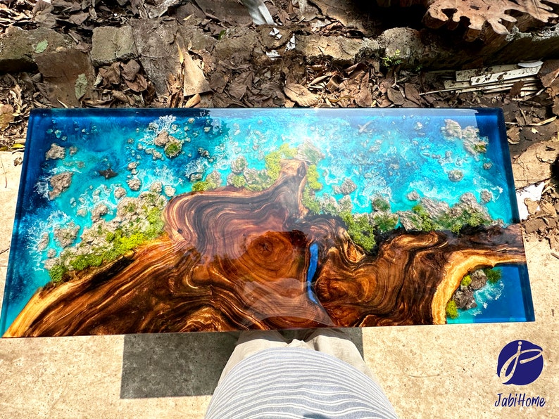 Puede incluir: Una mesa de centro rectangular con una base de madera oscura y una tapa de resina epoxi azul. La tapa de resina presenta un dise&ntilde;o de un oc&eacute;ano azul con olas blancas e islas verdes. La mesa mide aproximadamente 120 cm de largo y 60 cm de ancho.