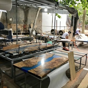 Puede incluir: Un taller con varias mesas de madera sin terminar con resina epoxi azul. Las mesas est&aacute;n sobre marcos de metal y se encuentran en diferentes etapas de finalizaci&oacute;n. Hay un gran ventilador en primer plano y una persona trabajando en una mesa en el fondo.