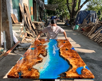 May include: A large, rectangular table with a blue epoxy river running through the center of the brown wood surface. The epoxy river has white waves and a blue-green color. The table is being held up by a person wearing a white shirt and a face mask.