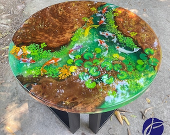 May include: A round wooden table top with a green epoxy resin finish featuring a design of koi fish swimming in a pond with lily pads. The table top is supported by a black wooden base.