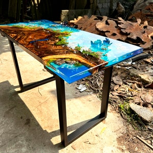 Puede incluir: Una mesa rectangular de madera con una tapa de resina epoxi azul y marr&oacute;n. La mesa tiene un marco de metal negro y est&aacute; sentada sobre una superficie de grava.