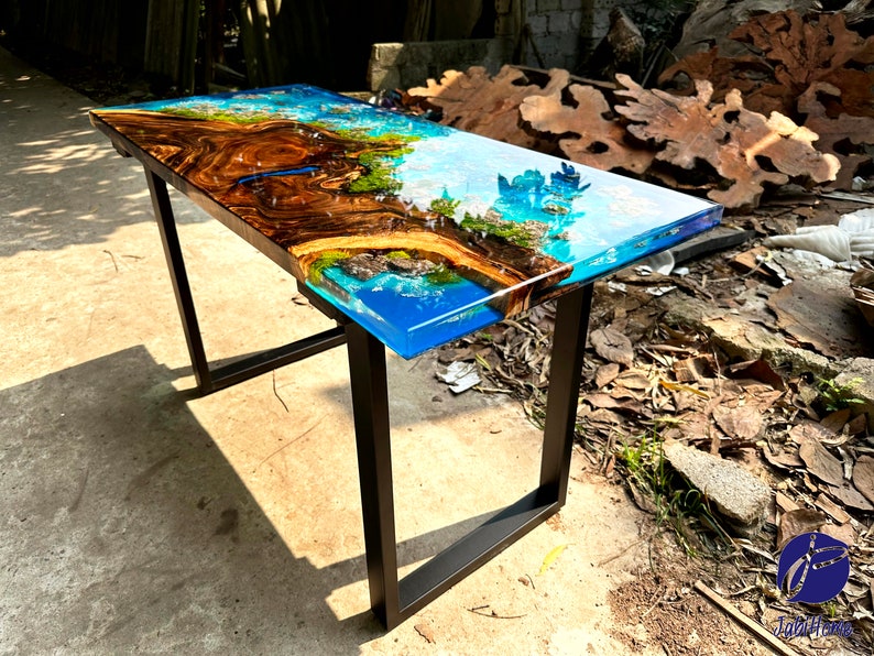 Puede incluir: Una mesa rectangular de madera con una tapa de resina epoxi azul y marr&oacute;n. La mesa tiene un marco de metal negro y est&aacute; sentada sobre una superficie de grava.