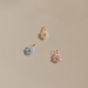 Op de afbeelding: Drie kleine bloemcharms gemaakt van kralen. De charms zijn lichtblauw, wit en roze. Elke charm heeft een gouden middelste kraal.