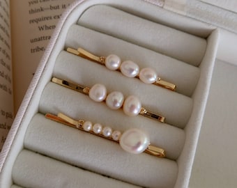 Puede incluir: Tres horquillas doradas con perlas de agua dulce de diferentes tamaños. Las perlas son blancas y tienen un brillo natural. Las horquillas están dispuestas en una caja de joyería blanca. Un accesorio para el cabello elegante.