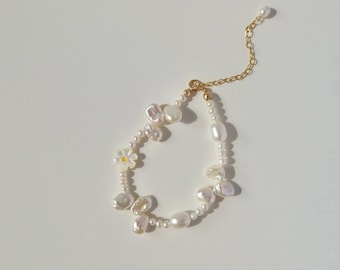 May include: A pearl bracelet with a mix of irregular and round pearls, and a small daisy charm. The bracelet has a gold-coloured chain and clasp. The background is white.