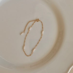 May include: A delicate gold chain bracelet with small white pearls. The bracelet is on a white plate.