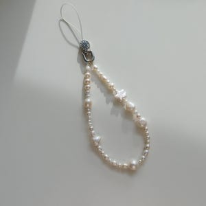 Può includere: Un charm per telefono con perle, con chiusura argentata e anello trasparente. Il charm presenta un mix di perle rotonde e irregolari, con dettagli a forma di stella. Le perle variano di dimensioni e hanno un colore bianco crema.
