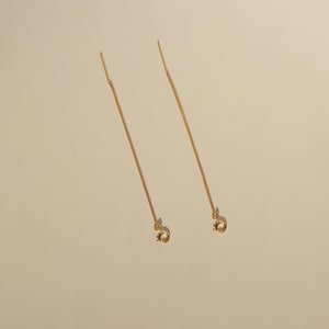 Pode incluir: Um par de brincos de corrente de ouro com um design simples e delicado. Os brincos são longos e pendentes, com um pequeno laço na ponta.