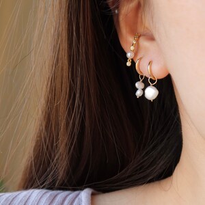 May include: A close-up of a person's ear with three gold earrings. The earrings are a small hoop, a pearl drop, and a curved bar with small pearls and diamonds.