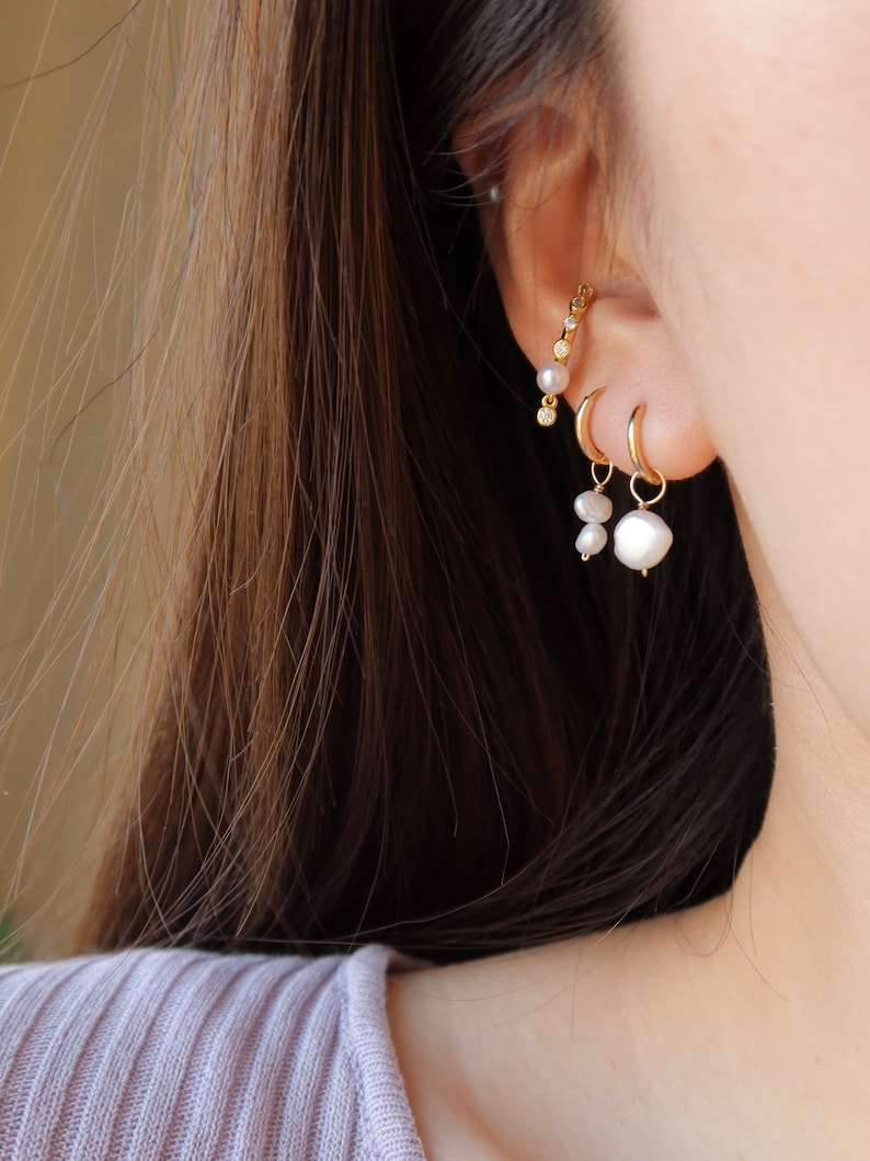 May include: A close-up of a person's ear with three gold earrings. The earrings are a small hoop, a pearl drop, and a curved bar with small pearls and diamonds.