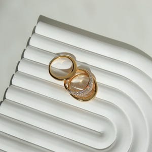 Può includere: Due anelli d'oro sono esposti su una superficie bianca e curva. Uno ha una fascia semplice, l'altro una fascia curva con piccole pietre preziose trasparenti. Gli anelli sono mostrati con luce naturale.
